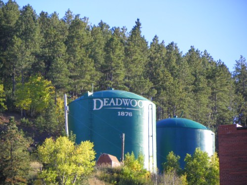 Two green water tanks labeled "DEADWOOD 1876" surrounded by trees and a clear blue sky.