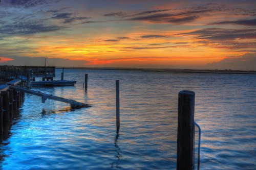 Sunset over calm waters, with a wooden dock and posts silhouetted against a colorful sky.