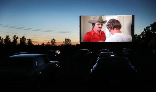 A drive-in movie screen shows a scene with two men, silhouetted cars in the foreground against a twilight sky.