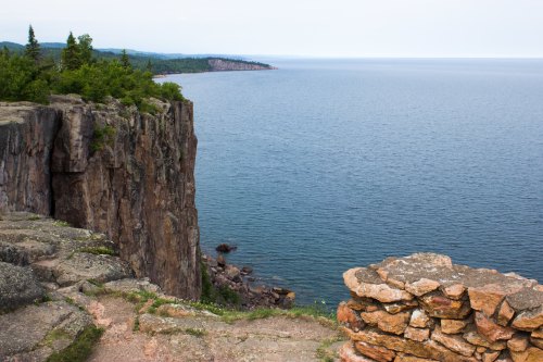 A rocky cliff overlooks a calm blue lake, with lush greenery and distant shoreline visible under a clear sky.