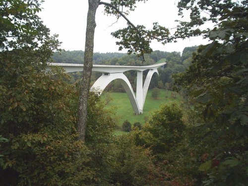 A modern white bridge with tall arches spans a green valley, surrounded by trees and hills.