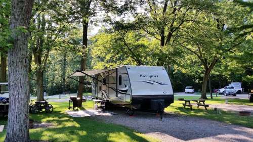 A travel trailer parked in a wooded campsite, surrounded by trees and picnic tables, with an awning extended.