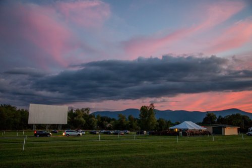 A drive-in movie theater at sunset, with a large screen, cars parked, and a tent under a colorful sky.