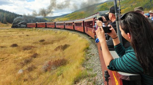 A steam train travels through a scenic landscape as passengers take photos from the train.