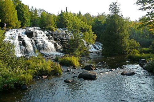 A serene waterfall cascades over rocks, surrounded by lush greenery and trees, reflecting a peaceful natural landscape.