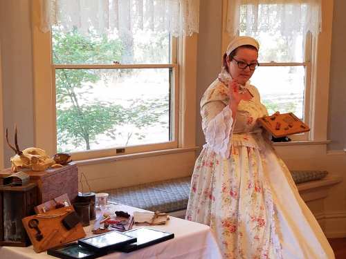 A woman in a floral dress gestures while standing by a table with historical artifacts and tools in a bright room.