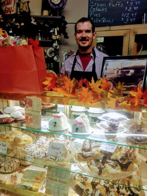 A smiling baker stands behind a display case filled with various pastries and desserts, decorated with autumn leaves.