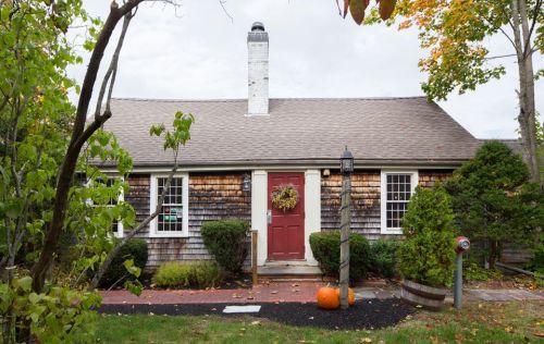 A charming cottage with a red door, surrounded by greenery and autumn decorations, including a pumpkin on the porch.