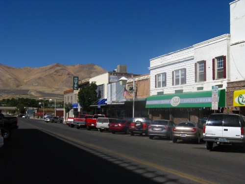 A small town street lined with shops and parked cars, with mountains in the background under a clear blue sky.