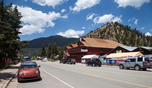 A scenic view of a small town street with parked cars, mountains in the background, and blue skies with clouds.