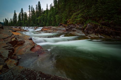 A serene river flows over rocky terrain, surrounded by lush green trees and a misty atmosphere.