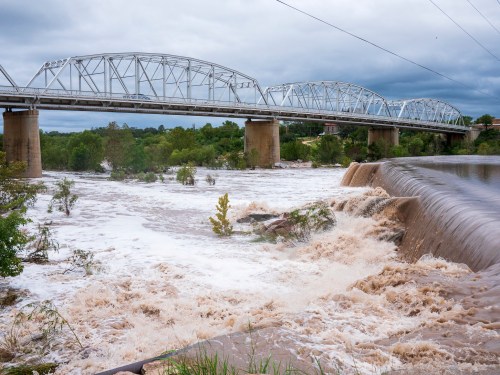 A bridge spans a river with turbulent, muddy water cascading over a dam, surrounded by greenery under a cloudy sky.