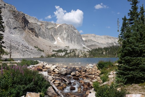 A serene mountain landscape featuring a clear lake, rocky shore, and lush greenery under a blue sky with clouds.