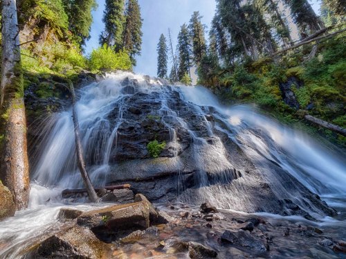 A cascading waterfall flows over rocky terrain, surrounded by lush green trees and a clear blue sky.