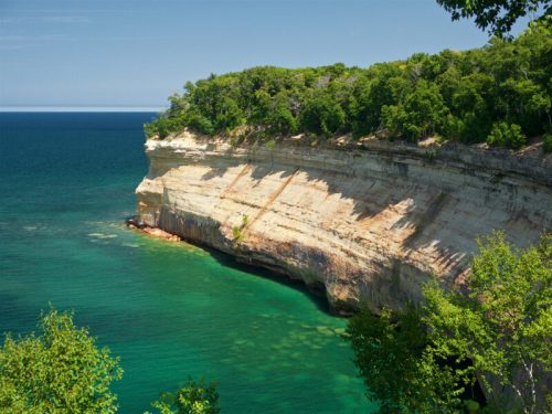 A scenic view of a rocky cliff overlooking a turquoise lake, surrounded by lush green trees under a clear blue sky.