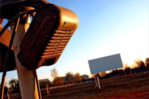 A close-up of a vintage drive-in speaker with a blank movie screen in the background during sunset.