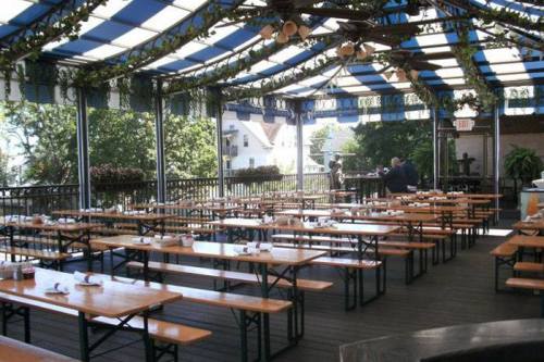 Outdoor dining area with wooden benches and tables, covered by a blue and white striped canopy, surrounded by greenery.