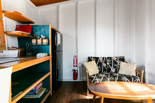 Cozy interior with a patterned couch, wooden shelves, a small table, and kitchen appliances in a bright, minimalist space.