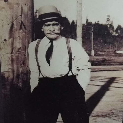 A vintage black-and-white photo of an older man in a hat and suspenders, standing by a wooden post.