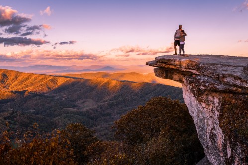A couple stands on a rocky ledge, overlooking a scenic mountain landscape at sunset, with vibrant clouds and valleys below.