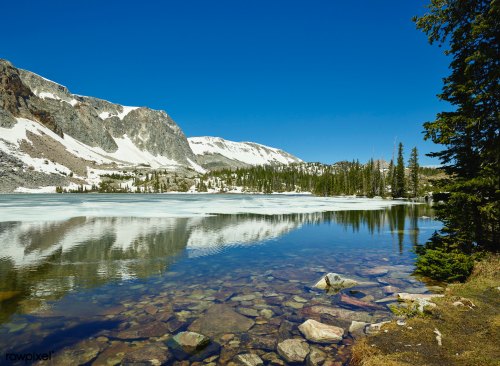 A serene mountain lake reflecting snow-capped peaks and clear blue skies, surrounded by evergreen trees.