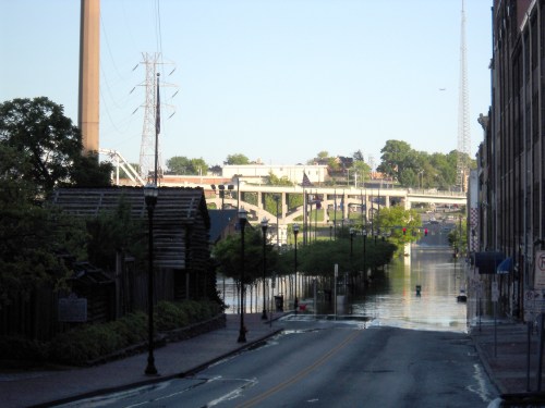 A flooded street with buildings on either side, power lines, and a bridge in the background under a clear sky.
