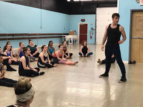 A dance instructor leads a class of students seated on the floor in a studio with blue walls.