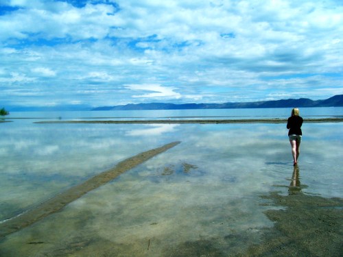 A person walks along a sandy shore, reflecting water and clouds under a blue sky with distant mountains.