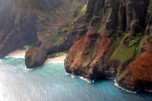 Aerial view of rugged cliffs meeting turquoise waters and a secluded beach. Lush greenery adorns the landscape.