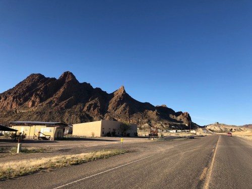 A deserted road leads to rugged mountains under a clear blue sky, with sparse buildings on the roadside.