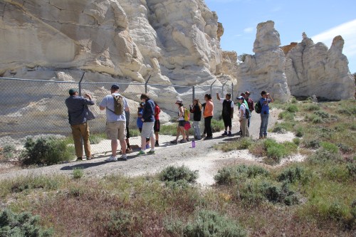 A group of people stands by a fence, observing rock formations in a desert landscape under a clear blue sky.