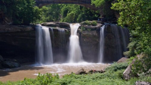 A scenic waterfall cascades over rocks, surrounded by lush greenery and a stone bridge in the background.