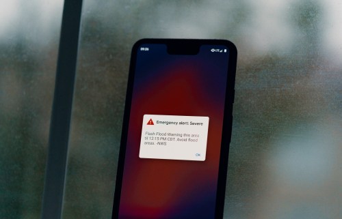 A smartphone displays a severe emergency alert for a flash flood warning, advising to avoid affected areas.