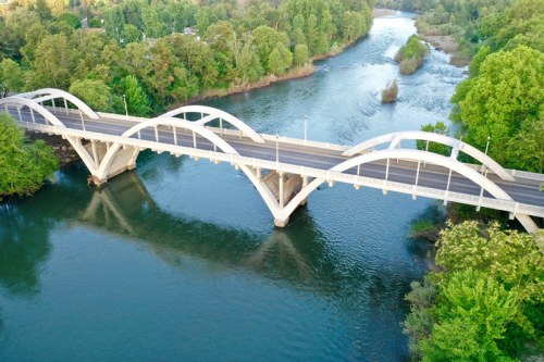 Aerial view of a white arch bridge spanning a river, surrounded by lush green trees and calm water.