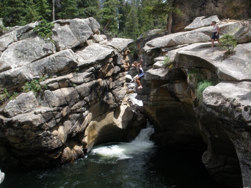 A person jumps from a rock ledge into a river surrounded by large boulders and trees in a natural setting.