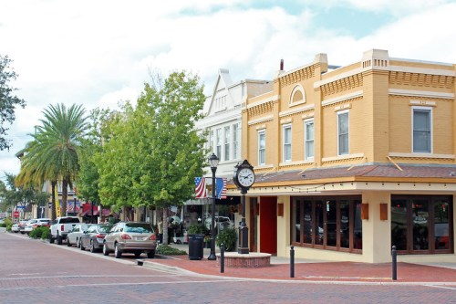 Charming downtown street with historic buildings, palm trees, and a clock, lined with parked cars and shops.