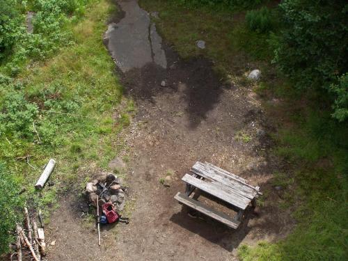A rustic picnic table in a grassy clearing, with a fire pit and scattered logs nearby.