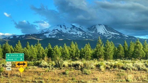 Snow-capped mountains rise above a forest, with road signs pointing to Dorris and Weed under a partly cloudy sky.