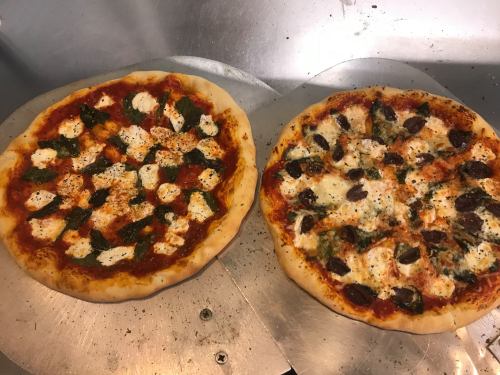 Two freshly baked pizzas on a metal surface: one with mozzarella and basil, the other with olives and cheese.