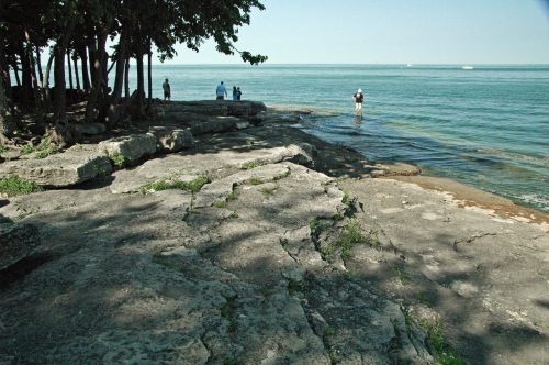 A rocky shoreline with trees, people standing by the water, and a calm lake in the background on a sunny day.