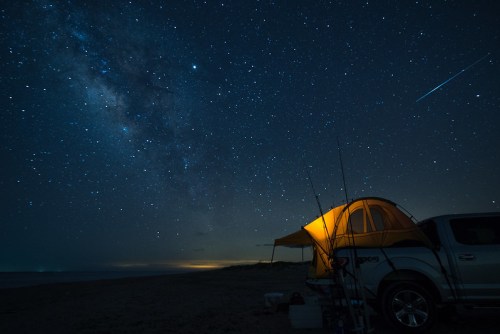 A tent glows under a starry night sky, with a truck nearby and fishing rods set up on a beach.