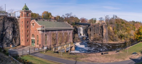 Historic building beside a waterfall, surrounded by trees and a clear sky, with a bridge in the background.