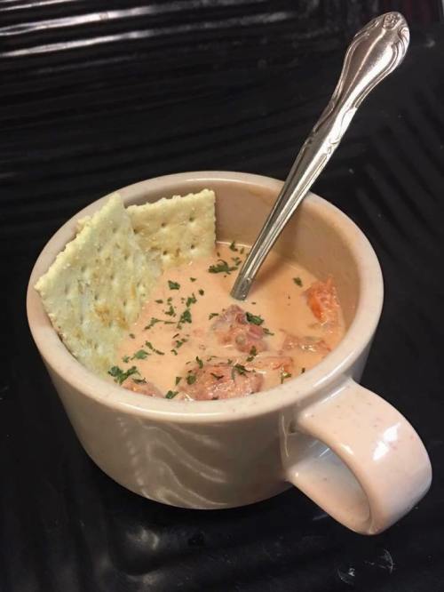 A cup of creamy soup with chunks of meat and vegetables, topped with two crackers and garnished with parsley.