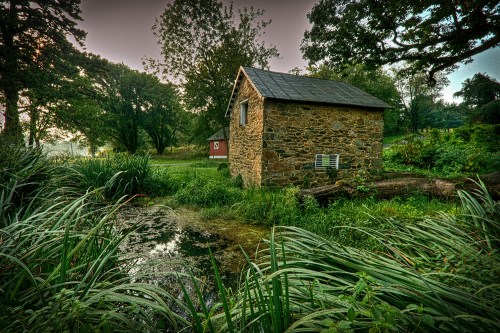 A rustic stone cottage surrounded by lush greenery and a small pond, set in a tranquil natural landscape.