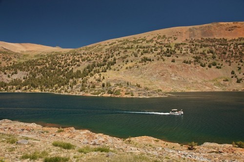 A boat glides across a serene lake surrounded by mountains and lush greenery under a clear blue sky.