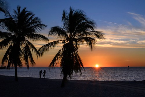 Silhouetted couple holding hands by the beach at sunset, with palm trees framing the colorful sky.
