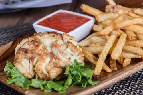 A wooden plate with a crab cake on lettuce, served with a side of fries and a small bowl of marinara sauce.
