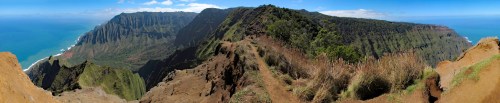 Panoramic view of a rugged coastline with cliffs, lush greenery, and a bright blue ocean under a clear sky.