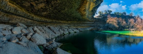 A serene landscape featuring a rocky overhang above a calm, greenish-blue body of water surrounded by trees.