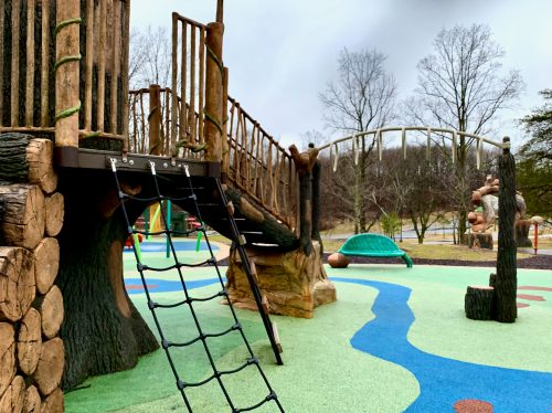 A playground with a wooden climbing structure, rope ladder, and colorful ground, surrounded by trees and a bridge in the background.